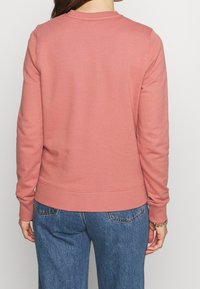 Różowa bluza z okrągłym dekoltem i prążkowanymi mankietami. Gładka tekstura, luźny krój i wykończenie w pasie. Połączona z dżinsami.