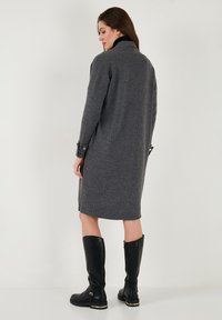 Robe en tricot côtelé gris avec un col et des manches longues. Associée à des bottes noires montantes avec des accents dorés. Vue arrière présentée.