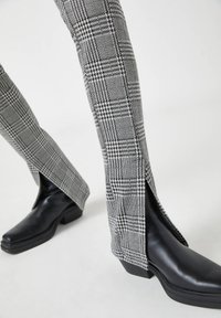 Pantalon gris à carreaux longueur jambe avec fentes à l’avant, porté sur des bottes à talons à bout carré noires sur un fond clair.