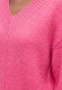Gros plan d'une personne portant un pull en maille rose vif à col en V montrant la texture détaillée du tissu et le col côtelé.