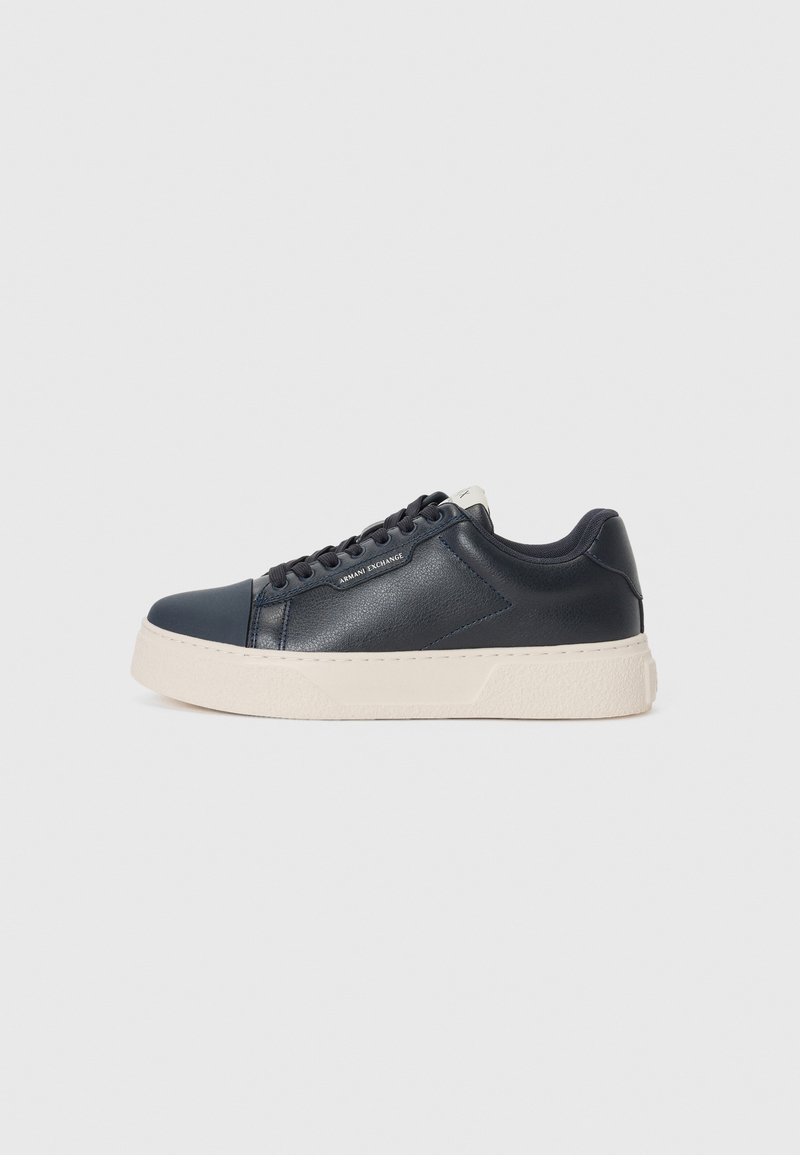 Marin lær sneaker med en teksturert finish, kontrasterende hvit gummisåle, rund tå og flate lisser, med subtil branding på siden.