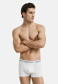 Weiße Baumwoll-Boxershorts mit einem marineblauen Logo-Bund, ausgestattet mit einer körpernahen Passform und flachen Nähten für zusätzlichen Komfort. Das Modell posiert mit verschränkten Armen.