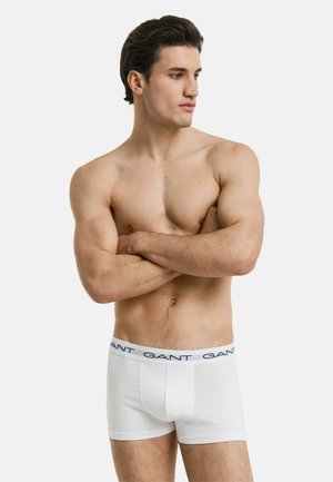 Weiße Baumwoll-Boxershorts mit einem marineblauen Logo-Bund, ausgestattet mit einer körpernahen Passform und flachen Nähten für zusätzlichen Komfort. Das Modell posiert mit verschränkten Armen.