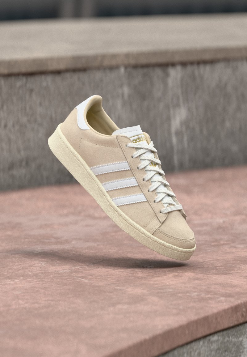 Bézs színű velúr tornacipő fehér csíkokkal, gumitalppal és lapos fűzőkkel. Arany logóval a cipőnyelven. Minimalista dizájnt mutat.