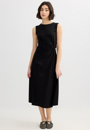 Mujer con vestido midi negro sin mangas con detalle de nudo lateral y zapatos Mary Jane negros de malla, de pie sobre un fondo liso y claro.