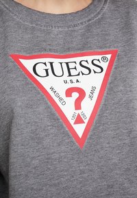 Harmaa neule, jossa on pyöreä kaula-auko ja suuri punainen kolmiomainen logo. Logossa on tekstit "GUESS U.S.A." ja kysymysmerkki, sekä teksti alapuolella.