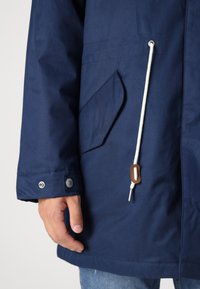 Marineblauwe waterdichte jas met een zak, witte trekkoord en bruine toggledetails. Geweven stof en knopenluiting bij de mouw.