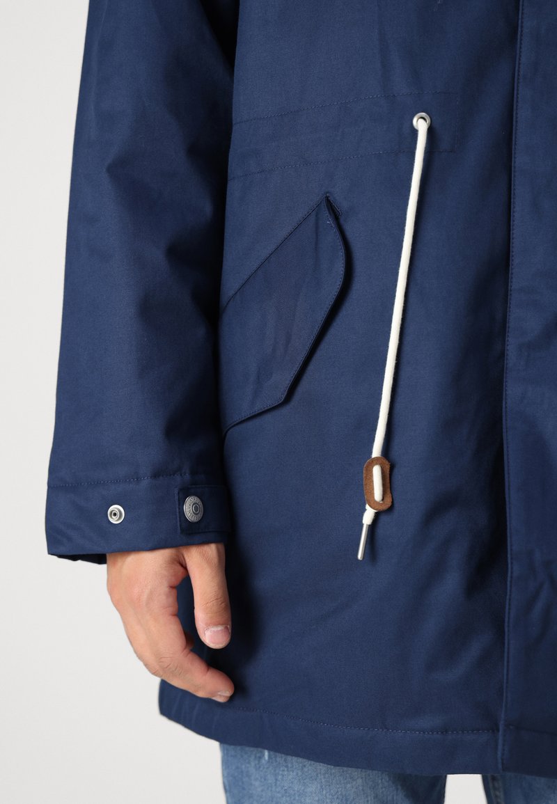 Giacca impermeabile blu navy con tasca, coulisse bianca e dettagli in legno marrone. Tessuto texturizzato e chiusura a bottone a pressione sul polsino.