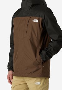 Personne portant une veste North Face marron et noire avec un logo sur la poitrine et un pantalon North Face beige avec un logo sur la cuisse.