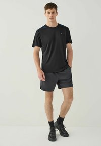 Sort atletisk t-shirt med blå skulderdetaljer, parret med grå shorts. Modellen har sorte sneakers og ankelsokker på.
