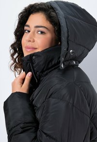 Chaqueta negra acolchada con cuello alto y capucha adjunta. Presenta botones a presión en el cuello y una apertura con cremallera. Textura suave y brillante.