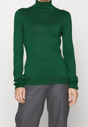 Persoon die een aansluitende groene coltrui en losse grijze broek draagt tegen een effen achtergrond.