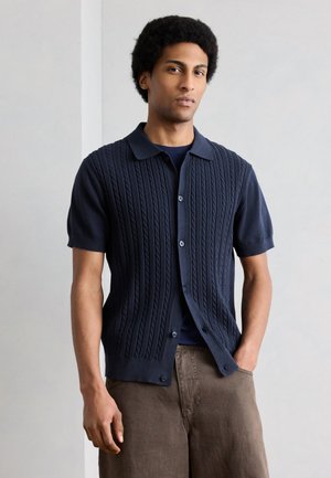 Mann med kort, krøllete hår iført en marineblå kabelstrikket kortermet cardigan over en mørk skjorte og brune bukser, stående mot en ensfarget bakgrunn.