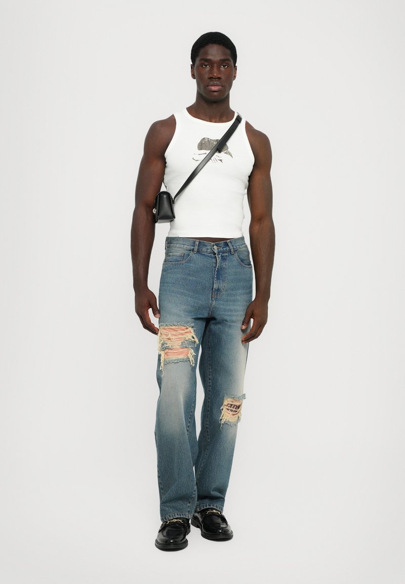 Weißes Tanktop mit Grafik, blaue, ausgebeulte Jeans mit großen Rissen und schwarze Schuhe. Kleine schwarze Umhängetasche über dem Oberkörper getragen.