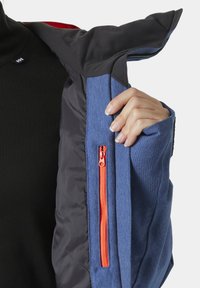 Blaue Jacke mit schwarzem Futter, ausgestattet mit einer orangefarbenen Reißverschluss-Tasche. Sichtbare Texturkontraste zwischen den Materialien, mit einem strukturierten Kragen-Design.