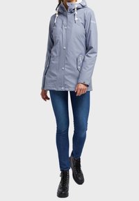 Veste imperméable gris clair avec capuche, cordons blancs, boutons-pression argentés et poches latérales, associée à un jean bleu foncé et des bottes noires.