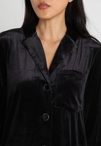 Czarny komplet piżamowy z aksamitu, z kołnierzykiem, kieszonką z przodu i zapięciem na guziki. Materiał ma miękką, gładką teksturę.