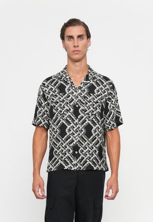 Young man wearing black and white patterned short-sleeve button-up shirt and black cargo pants, standing against a plain white background.