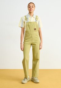 Salopette verde chiaro con spalline regolabili, tasca frontale e patch con logo, abbinata a una maglietta a righe giallo chiaro e blu. Sneakers beige.