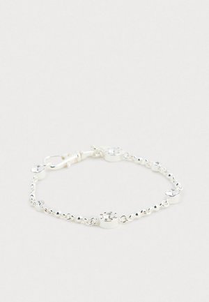 Srebrna verižica za zapestje s okroglimi prozornimi kristali in majhnim graviranim amuletom, postavljena na enostavno svetlo ozadje.