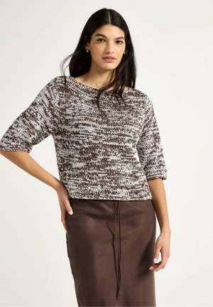 Kvinde med langt mørkt hår iført en brun og hvid strikket sweater og en brun nederdel, poserer med den ene hånd på hoften.