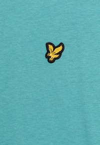 Tyrkysové bavlněné tričko s malým černým a zlatým vyšitým logem ptáka na levé části hrudi. Hladká textura, volný styl.