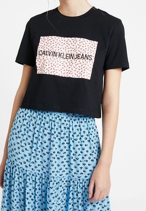 Černé krátké tričko s obdélníkovou záplatou s květinovým vzorem a textem "CALVIN KLEIN JEANS". Kombinováno s modrou květinovou maxi sukní.