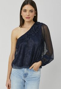 Haut asymétrique bleu marine avec détails en volants, manches longues transparentes et tissu texturé. Associé à un jean en denim bleu clair.