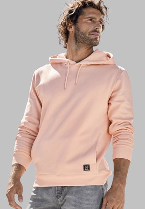 Peach cotton hoodie with a kangaroo pocket, drawstring hood, ribbed cuffs, and a small black logo patch at the hem. Gray background.
