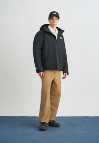 Zwarte waterdichte jas met capuchon en ritssluitingen, gecombineerd met beige wijde broekspijpen en zwarte schoenen. Model draagt een zwarte pet.