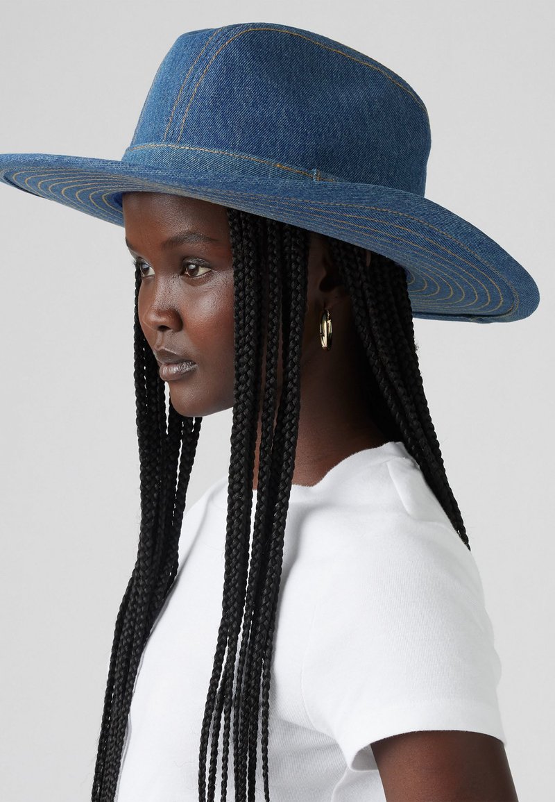 Cappello in denim a tesa larga con cuciture gialle a contrasto, caratterizzato da una corona rigida e una texture liscia. Indossato con una camicia bianca semplice.