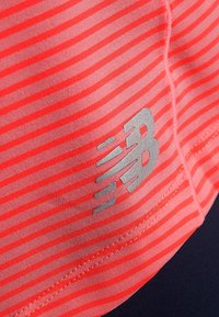 Červené a růžové pruhované látky s reflexním stříbrným logem "NB", hladkou texturou a přiléhavým designem, ideální pro sportovní oblečení.