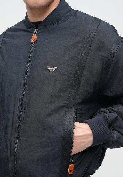 Čierna bomber bunda so zapínaním na zips, rebrovanými manžetami a odznakom s logom. Vyznačuje sa textúrovanou tkaninou a kontrastnými bočnými popruhmi. Ozdobný ťahač zipsu.