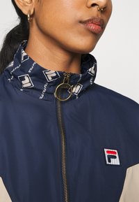 Giubbotto a vento navy con colletto a fantasia, zip dorata e patch logo FILA; presenta dettagli beige a contrasto sulle spalle.