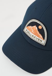 Gorra de béisbol azul marino con visera curva. Presenta un logo de montaña bordado con un sol naranja. Está hecha de un tejido suave con costuras visibles.