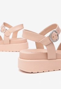 Beige platform sandals feature crossed straps, a circular buckle detail, and a textured sole for grip and comfort.