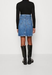 Person som bär en svart långärmad topp, blå denimkjol med bakfickor och svarta knähöga stövlar, står mot en plain vit bakgrund.