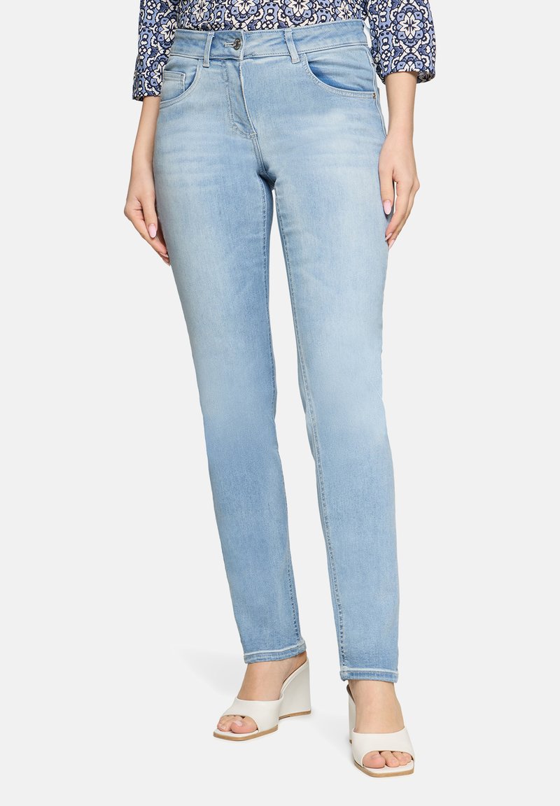 Jean en denim bleu clair à coupe slim, cinq poches, et effet légèrement délavé. Associé à des sandales blanches à talons.