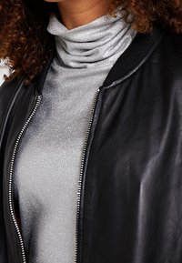Černá kožená bomber bunda s třpytivým stříbrným rolákem. Bunda má zapínání na zip a žebrovaný límec. Viditelná lesklá texturovaná tkanina.