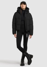 Juodas pūkinis švarkas su nusidėvimu kiseniu ir užtrauktuku, derinamas su juodomis prigludusiomis legging'ais ir koviniais batais. Paprastas dizainas, be raštų.