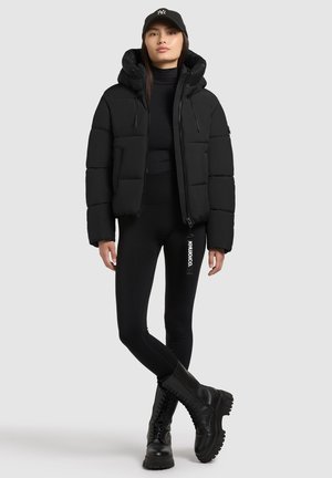 Svart puffjacka med avtagbar huva och dragkedjestängning, kombinerad med svarta stretchleggings och kängor. Enkel design, utan mönster.