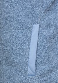 Jasnoniebieska tkanina polarowa o teksturowej, bąbelkowej powierzchni oraz gładkiej, płaskiej części. Zawiera boczną kieszeń z pasującego materiału.