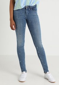 Snygga blå denimjeans i slim fit med subtil blommönstring, med medelhög midja och ankellång passform, kombinerade med vita sneakers.