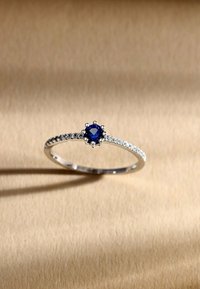 Silver ring med en central blå ädelsten, omgiven av mindre diamantaccenter längs en liten ring. Slät och polerad yta.