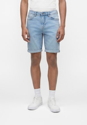 Uomo che indossa pantaloncini di jeans blu chiaro con orlo arrotolato, calzini bianchi e scarpe da ginnastica bianche, in piedi davanti a uno sfondo bianco semplice.