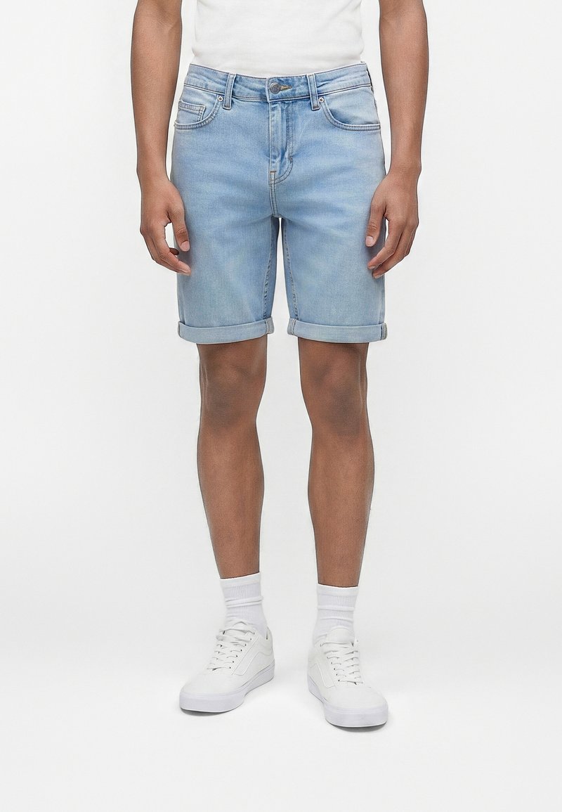 Άνδρας που φοράει ανοιχτό μπλε τζιν σορτς με γυρισμένα μπατζάκια, λευκές κάλτσες και λευκά αθλητικά παπούτσια, στέκεται μπροστά σε μια απλή λευκή επιφάνεια.