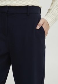 Navy pants made of a smooth, stretchy fabric feature a straight cut and side pockets, with a waistband and fingers resting in the pocket.