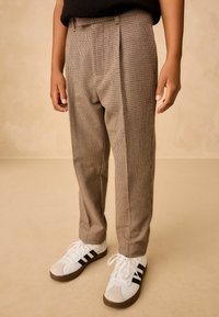 Brown checkered trousers with a relaxed fit, featuring a pleated front and side pockets, paired with white and black athletic shoes.