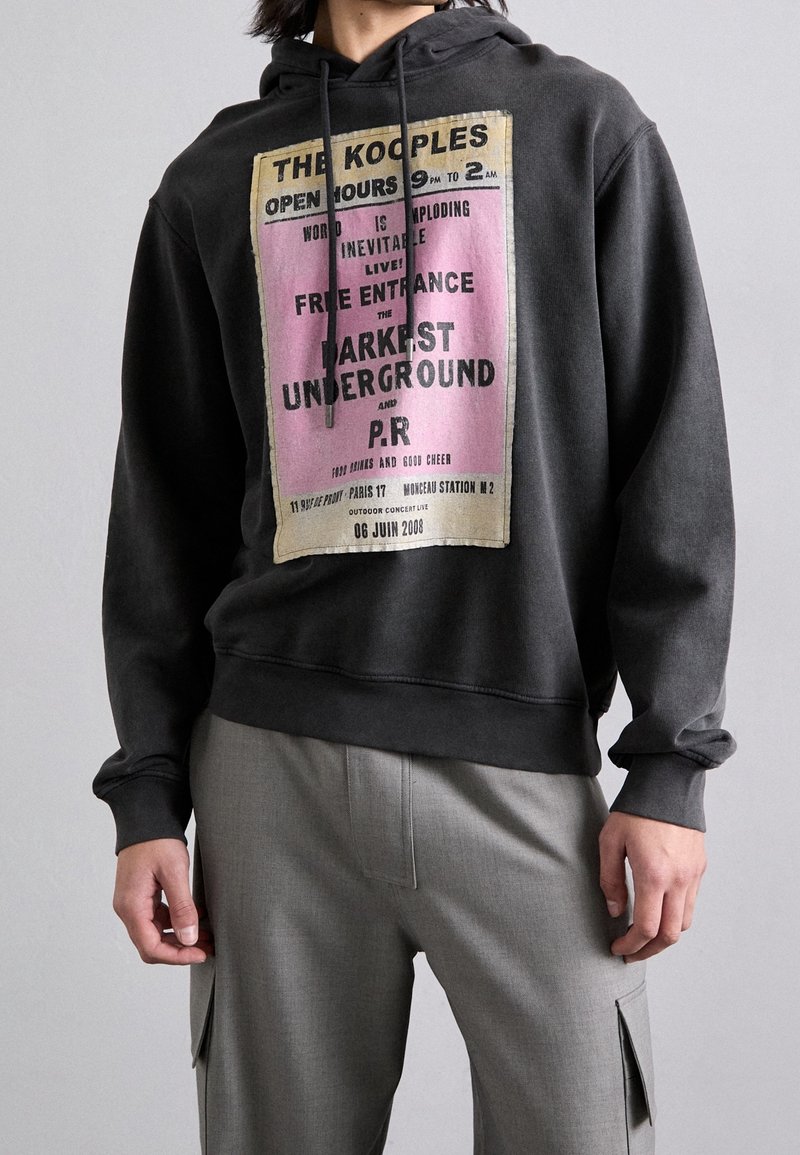 Felpa nera con grafica di pubblicità di concerti vintage in vari colori, dotata di lacci e vestibilità rilassata. Pantaloni grigi visibili sotto.