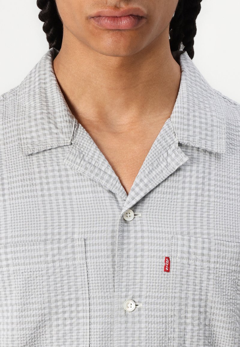 Chemise à manches courtes à carreaux gris avec un col, dotée d'une poche plaquée et de boutons blancs. Étiquette rouge Levi's sur la poche gauche.